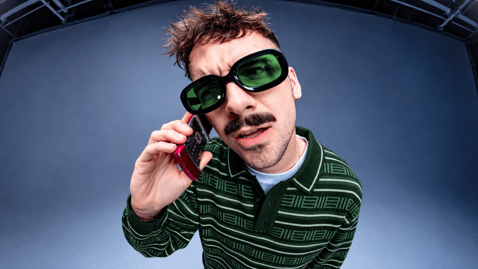 A creator in green-tinted sunglasses and a striped polo leaning into a fisheye lens while talking on a flip phone.