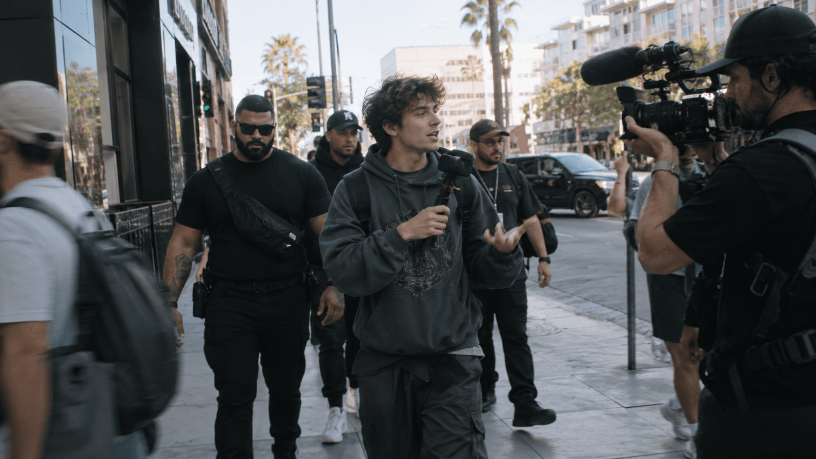 A streamer walking down a city street holding a camera rig, followed by security and filmed by a camera operator.